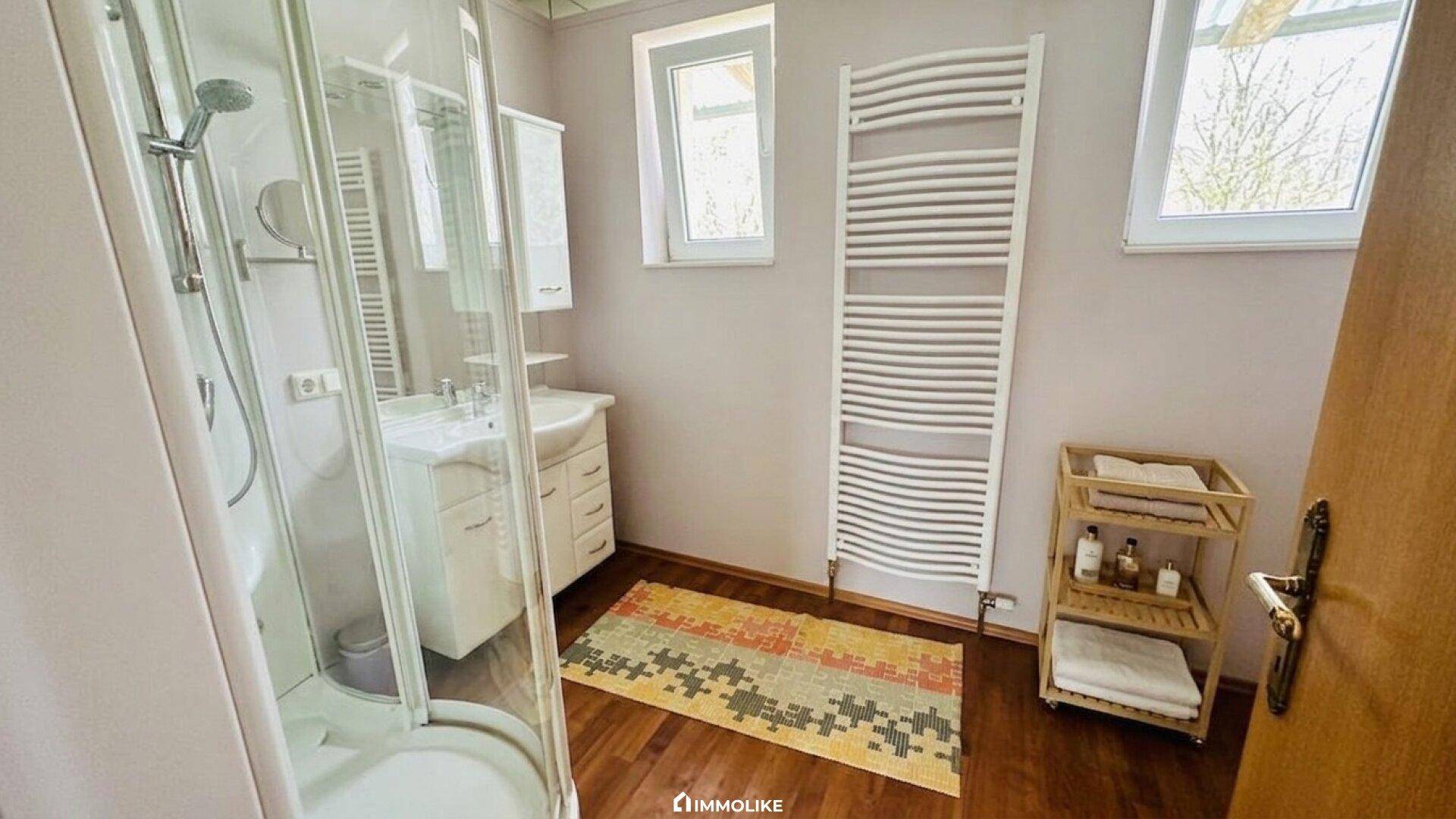 Helles Badezimmer mit Dusche, Waschbecken, beheiztem Handtuchhalter und zwei Fenstern.