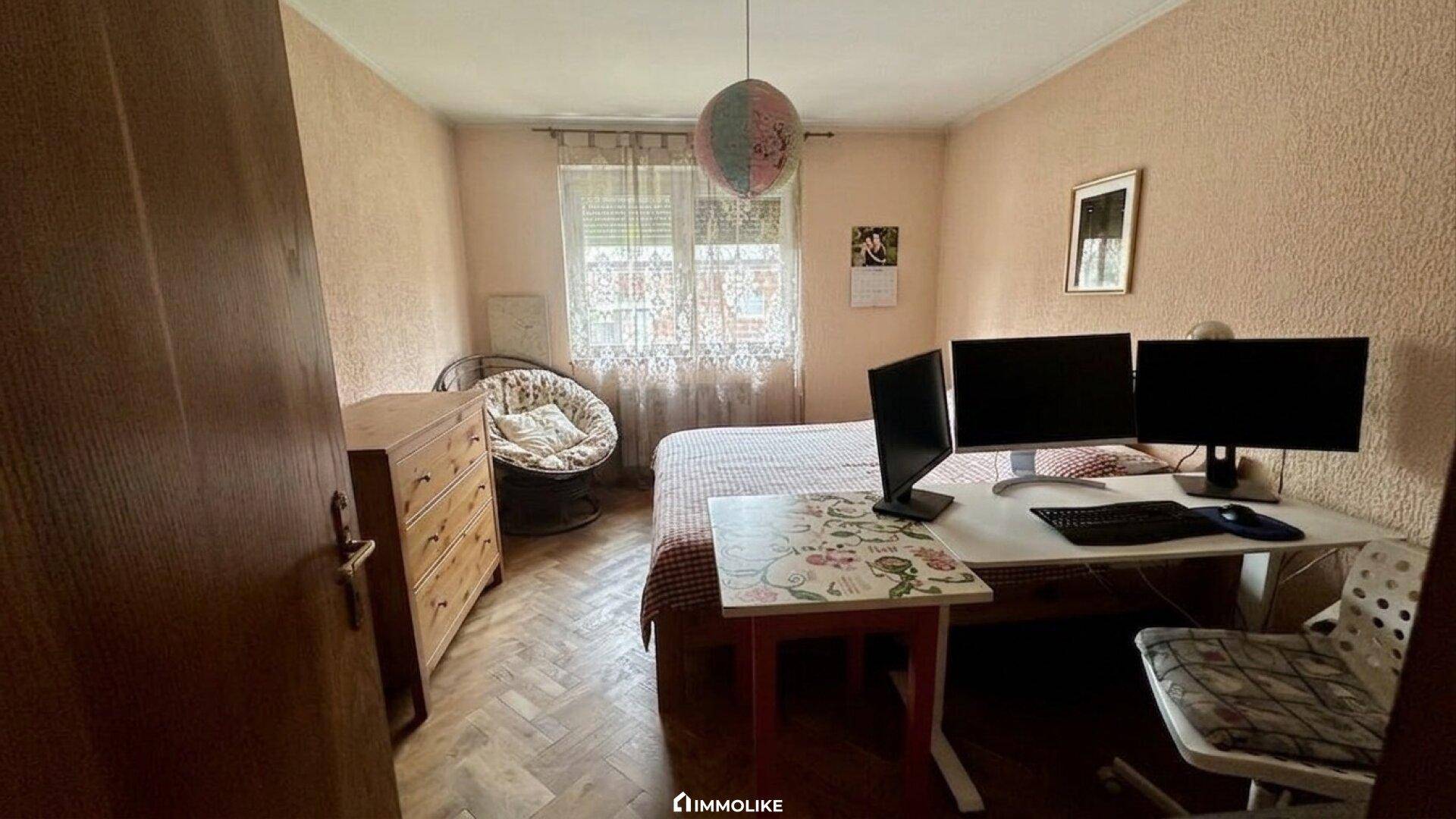 Schlafzimmer mit Parkettboden, Schreibtisch, zwei Monitoren und Fenster.