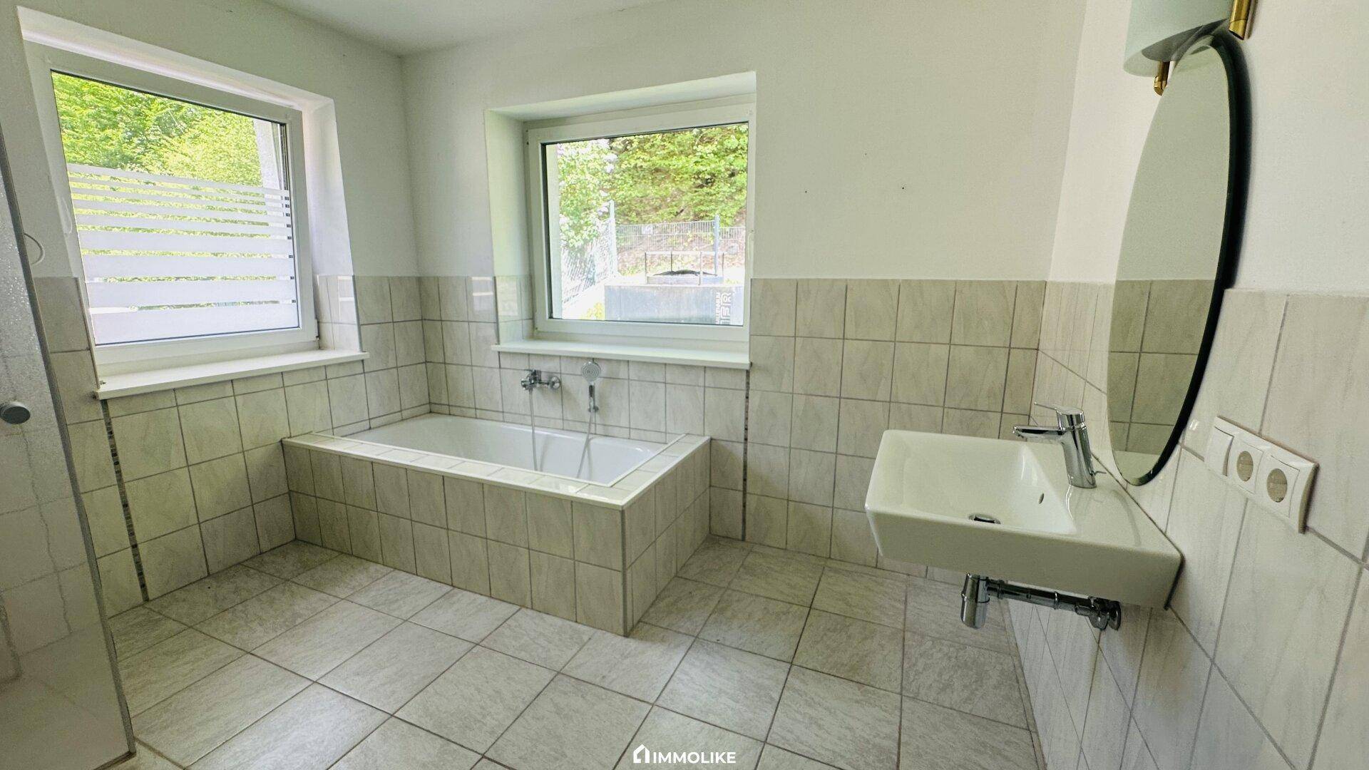 Helles Badezimmer mit Badewanne, Waschbecken, Spiegel und zwei Fenstern für Tageslicht.