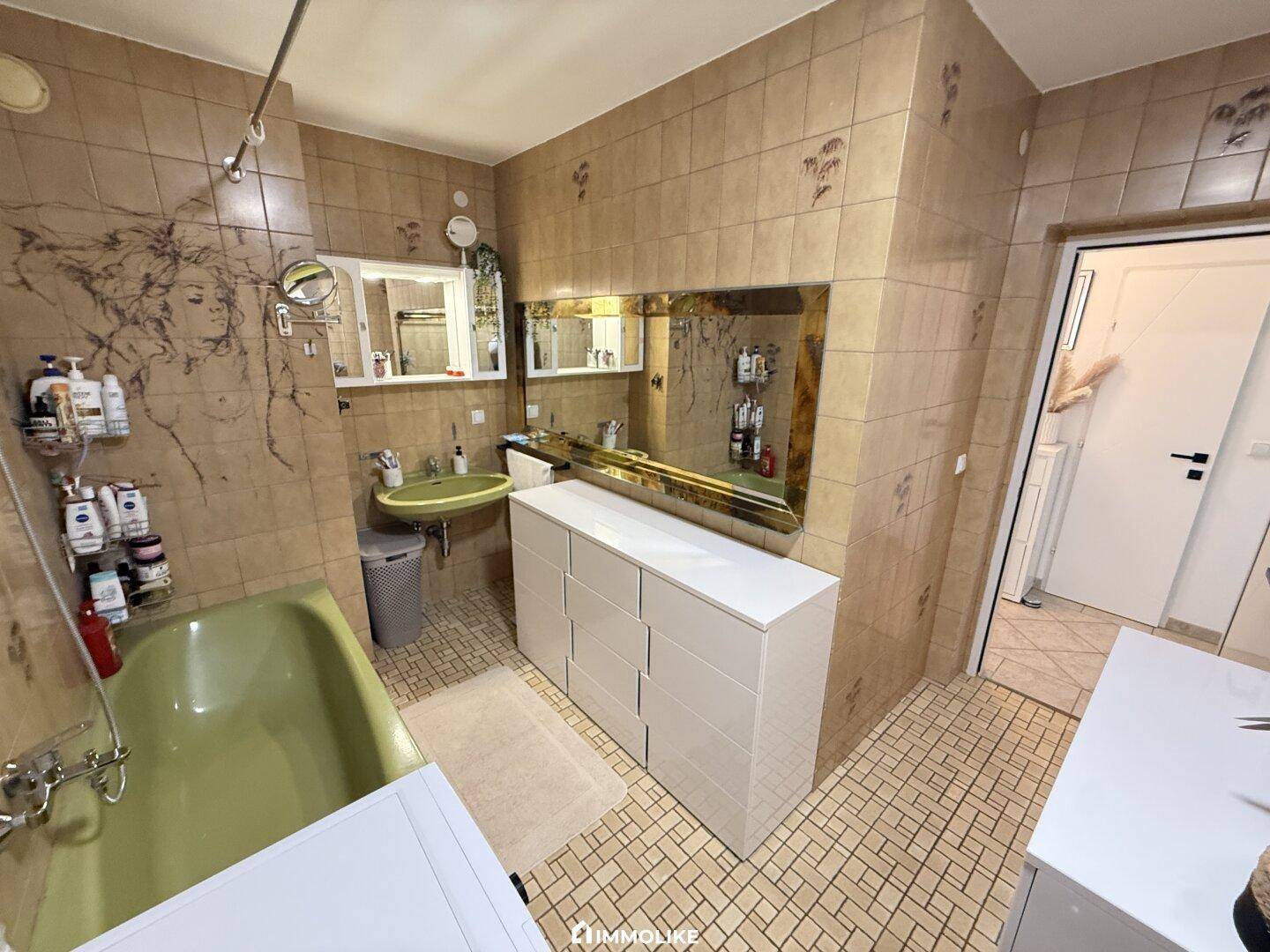 Badezimmer mit Badewanne, Waschbecken und großem Spiegel, gefliest in Brauntönen.