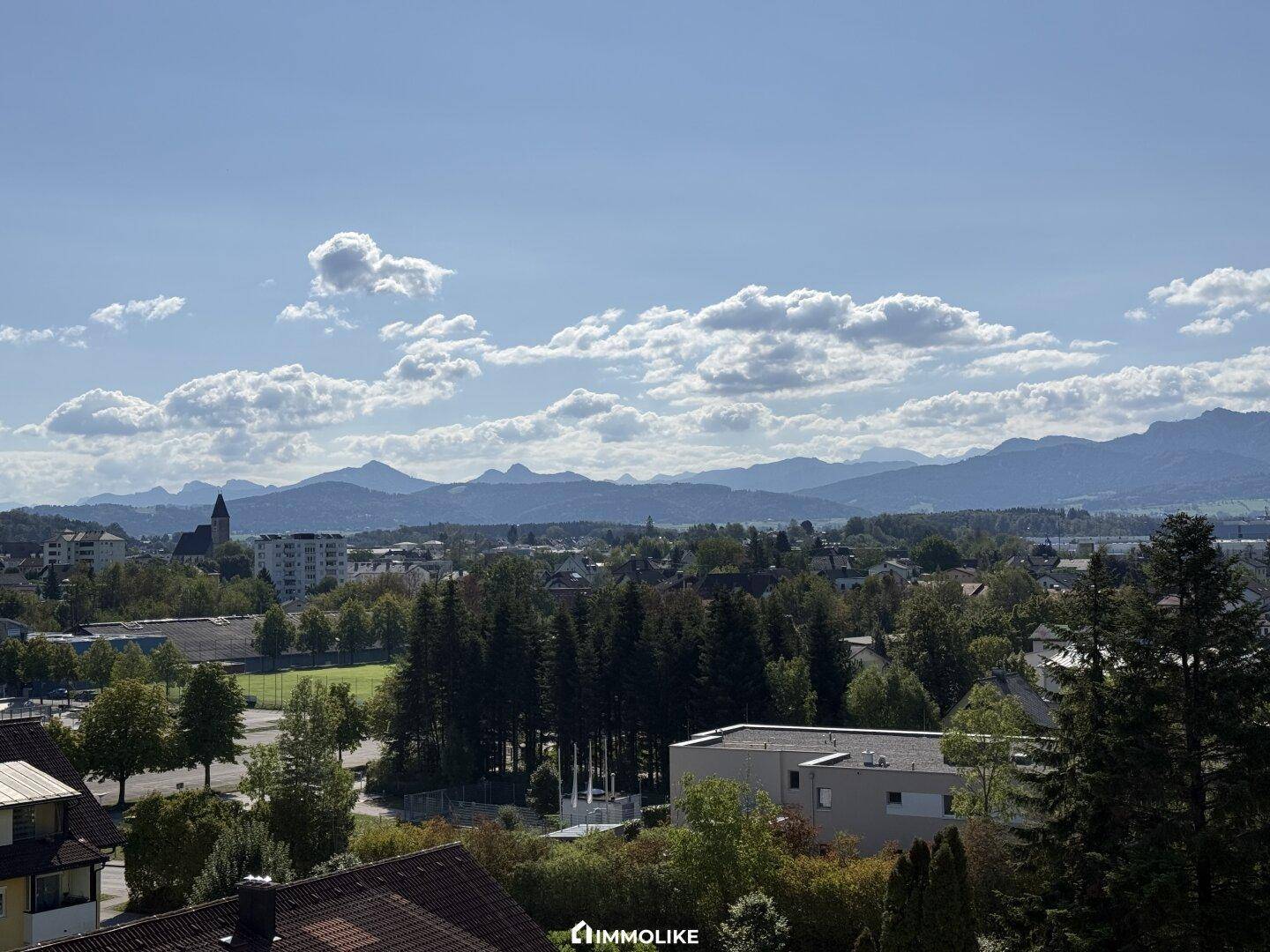 Panoramablick auf die Stadt, grüne Landschaften und die umliegenden Berge unter blauem Himmel.