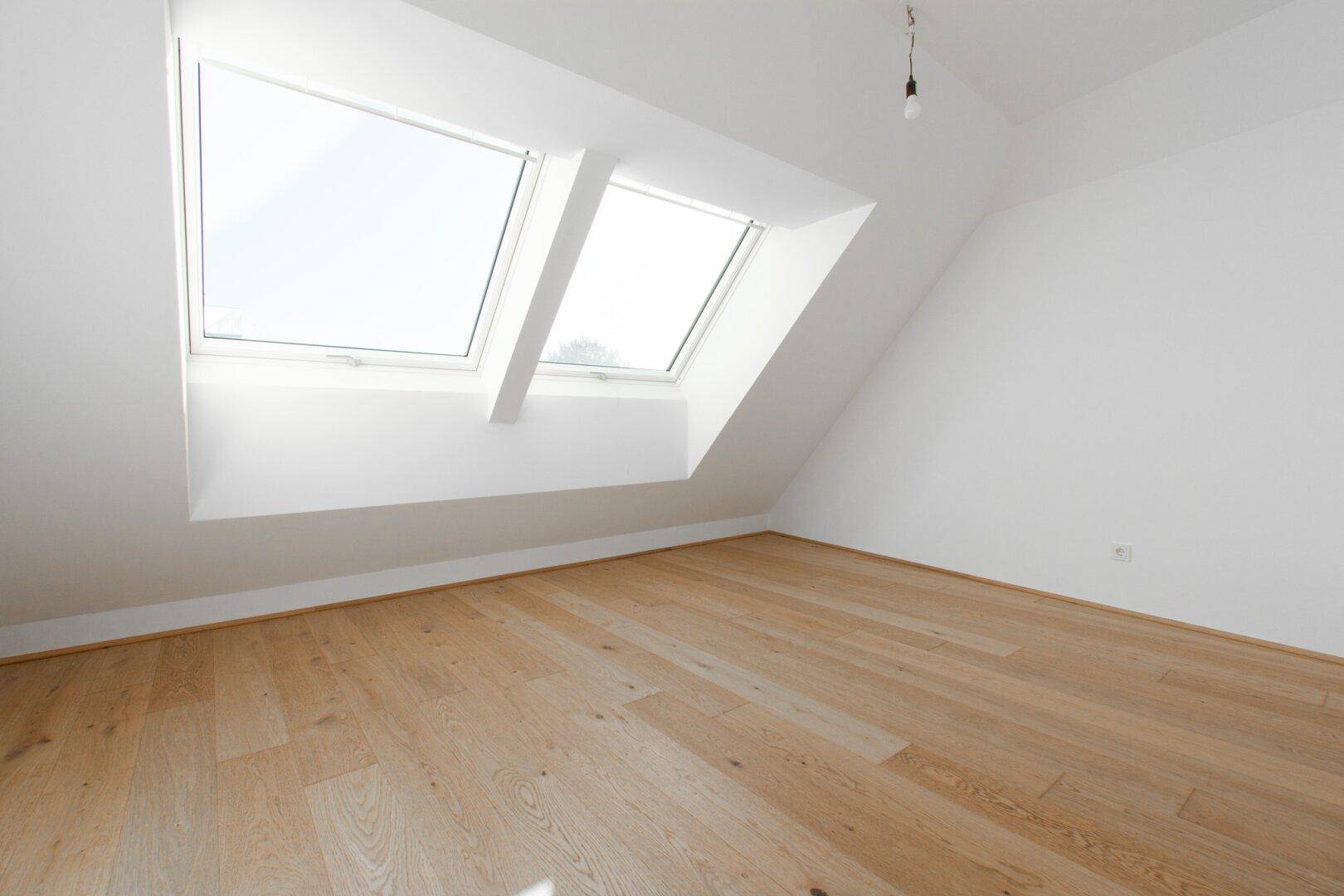 Helles Dachzimmer mit zwei großen Dachfenstern und schrägen Wänden, ideal als Schlafzimmer.