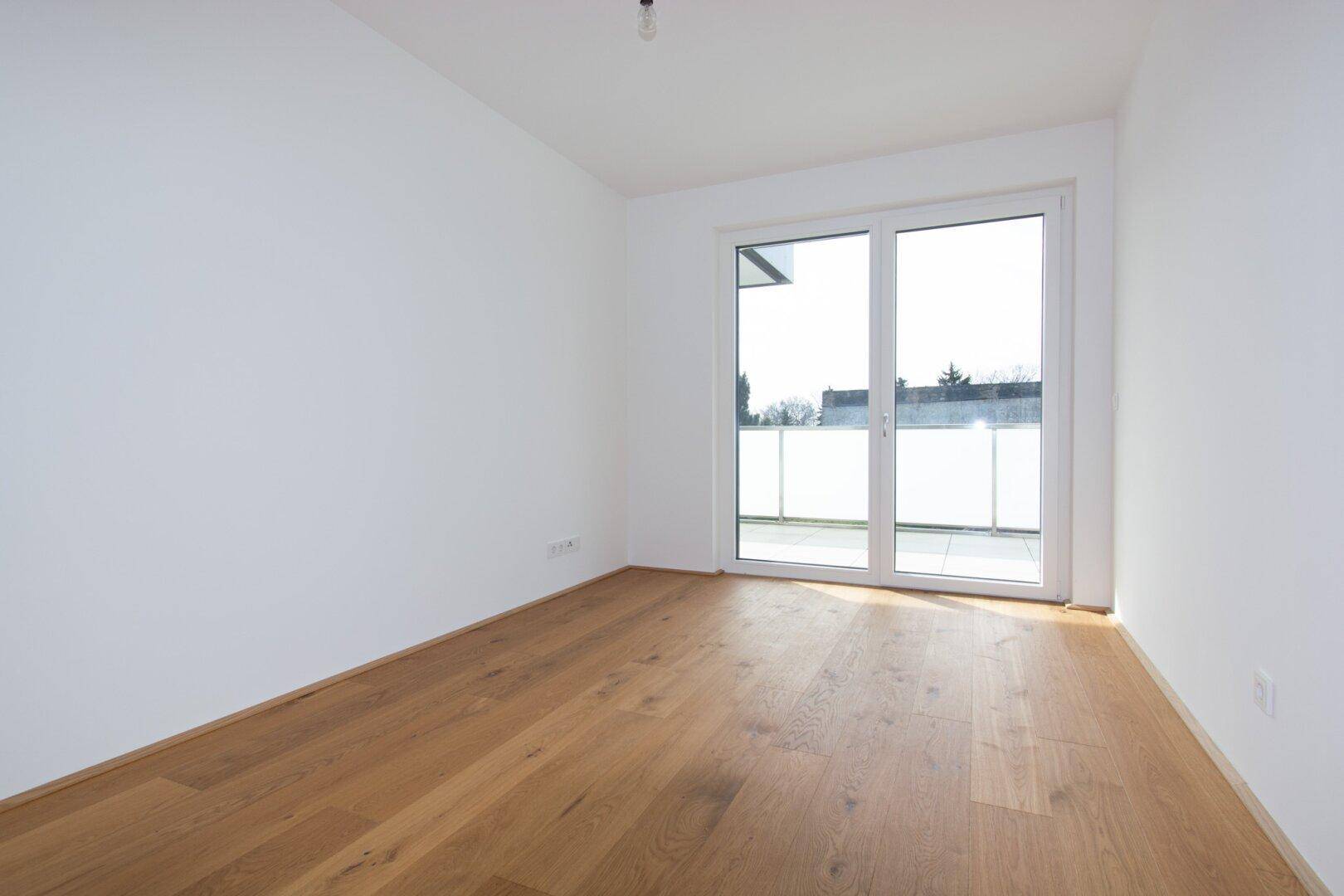 Heller Raum mit Holzboden und großen Fenstern, die Zugang zu einem Balkon bieten.