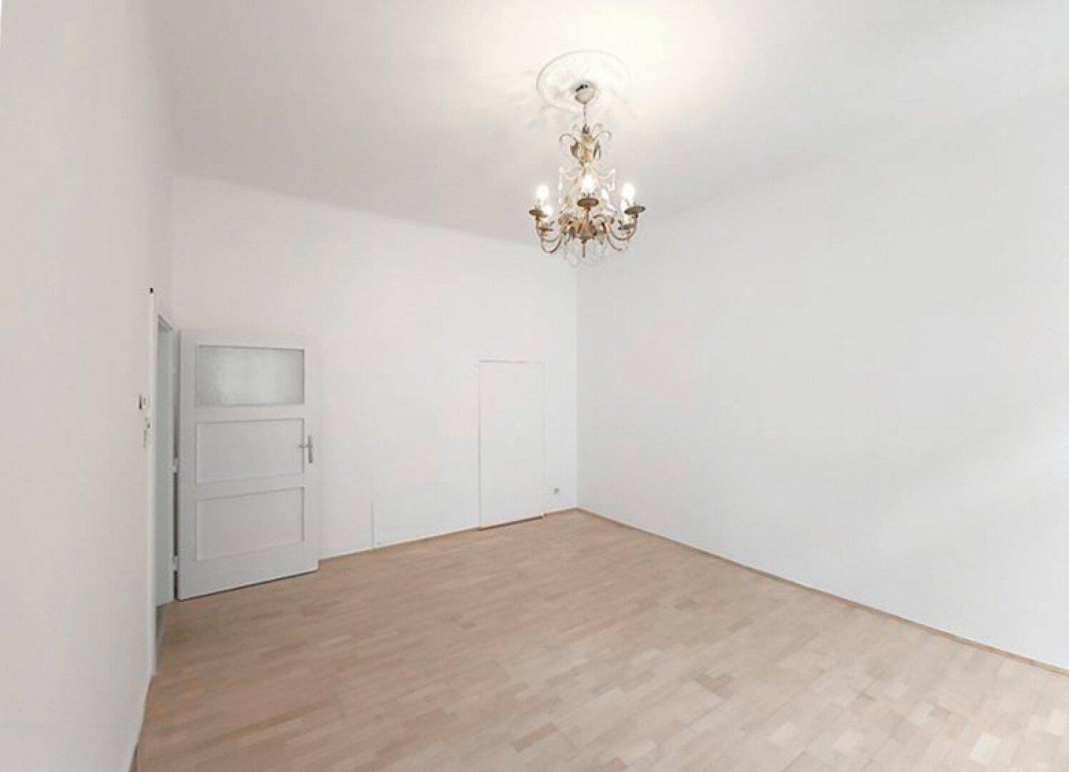 Geräumiger, heller Raum mit Parkettboden, klassischem Kronleuchter und einer weißen Tür im Altbau-Stil.