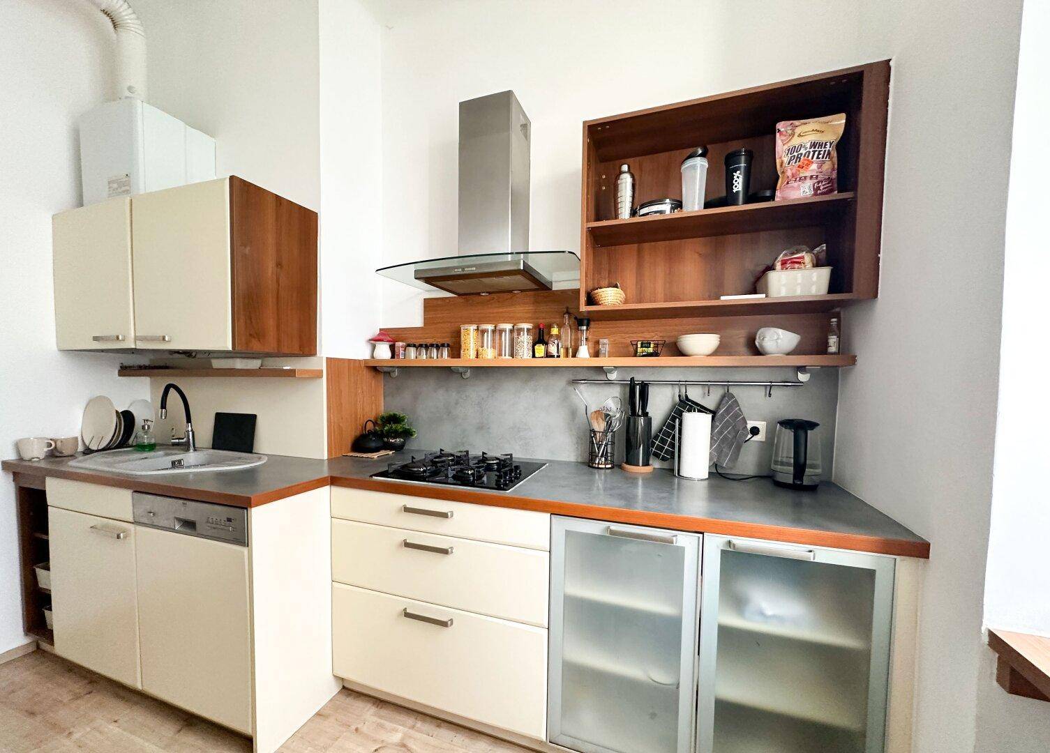 Funktionale Küche mit Gasherd, Holzregalen, hellen Schränken und einem sichtbaren Warmwasserboiler.