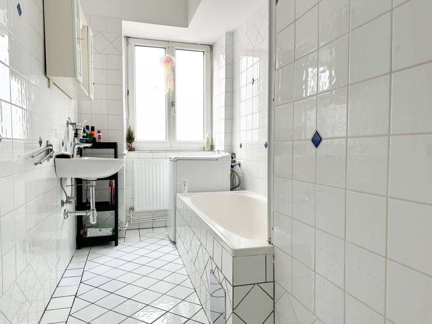 Helles Badezimmer mit weißen Fliesen, Badewanne und Fenster für natürliche Belüftung und Tageslicht.