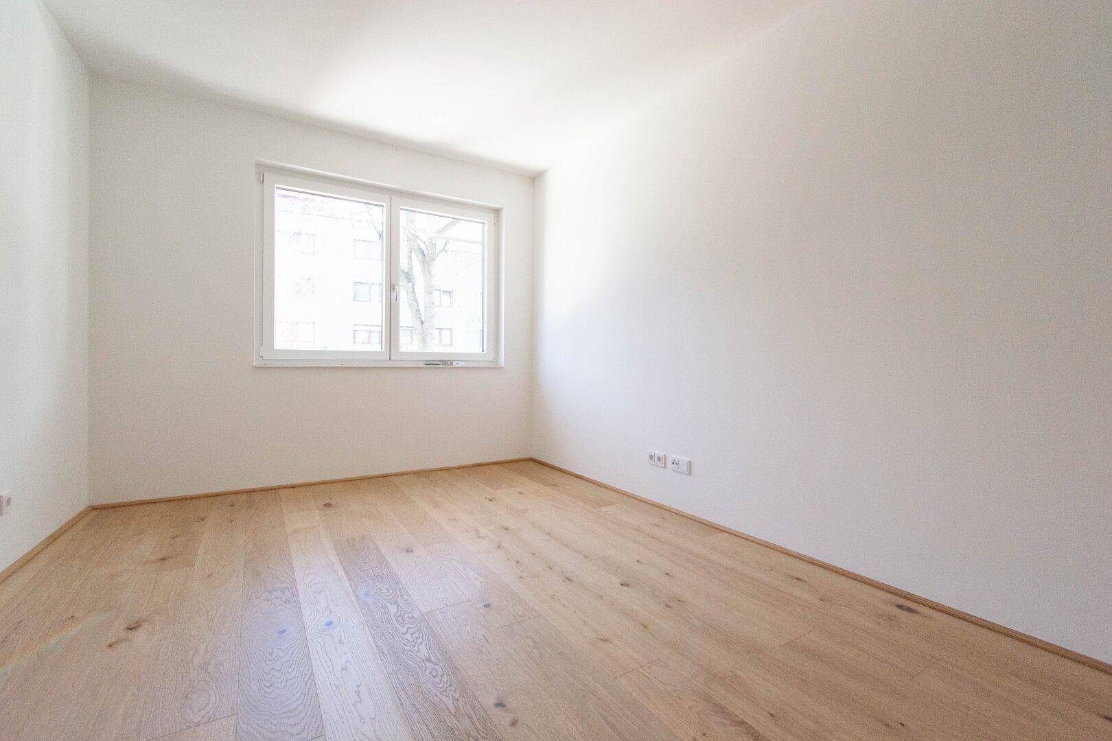 Helles Schlafzimmer mit Parkettboden und einem Fenster, das viel Tageslicht hereinlässt.
