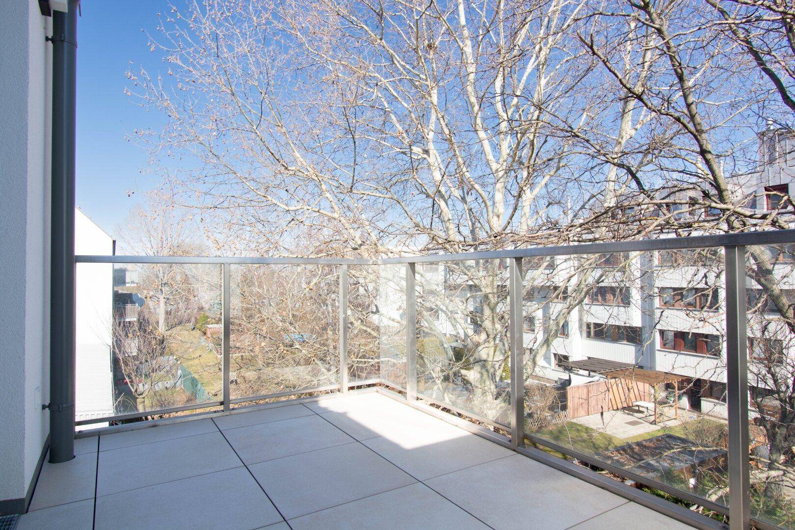 Heller Balkon mit Fliesen und Glasgeländer, umgeben von Bäumen und Gebäuden.