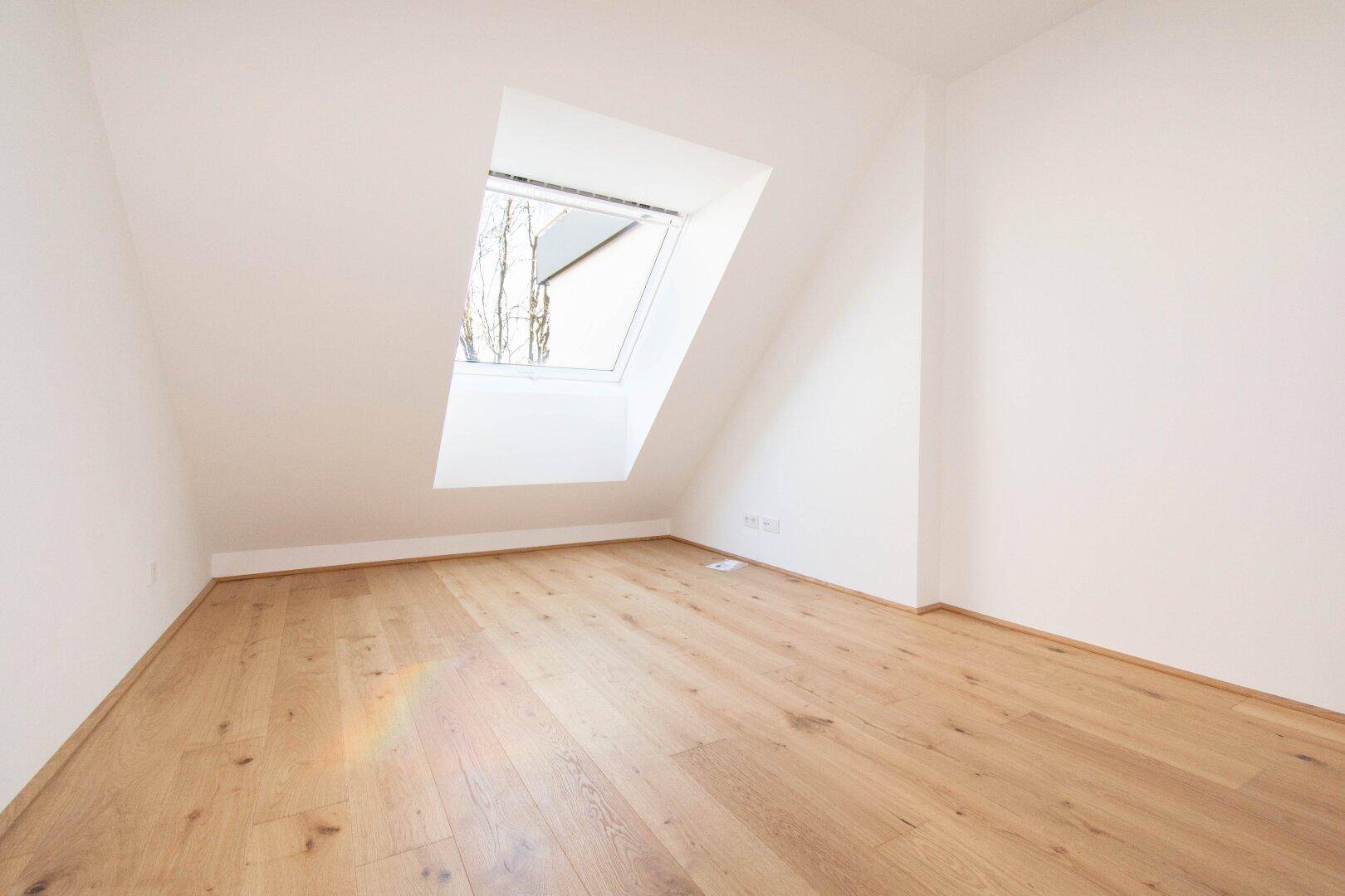 Dachzimmer mit schrägen Wänden, hellem Holzboden und einem großen Dachfenster.