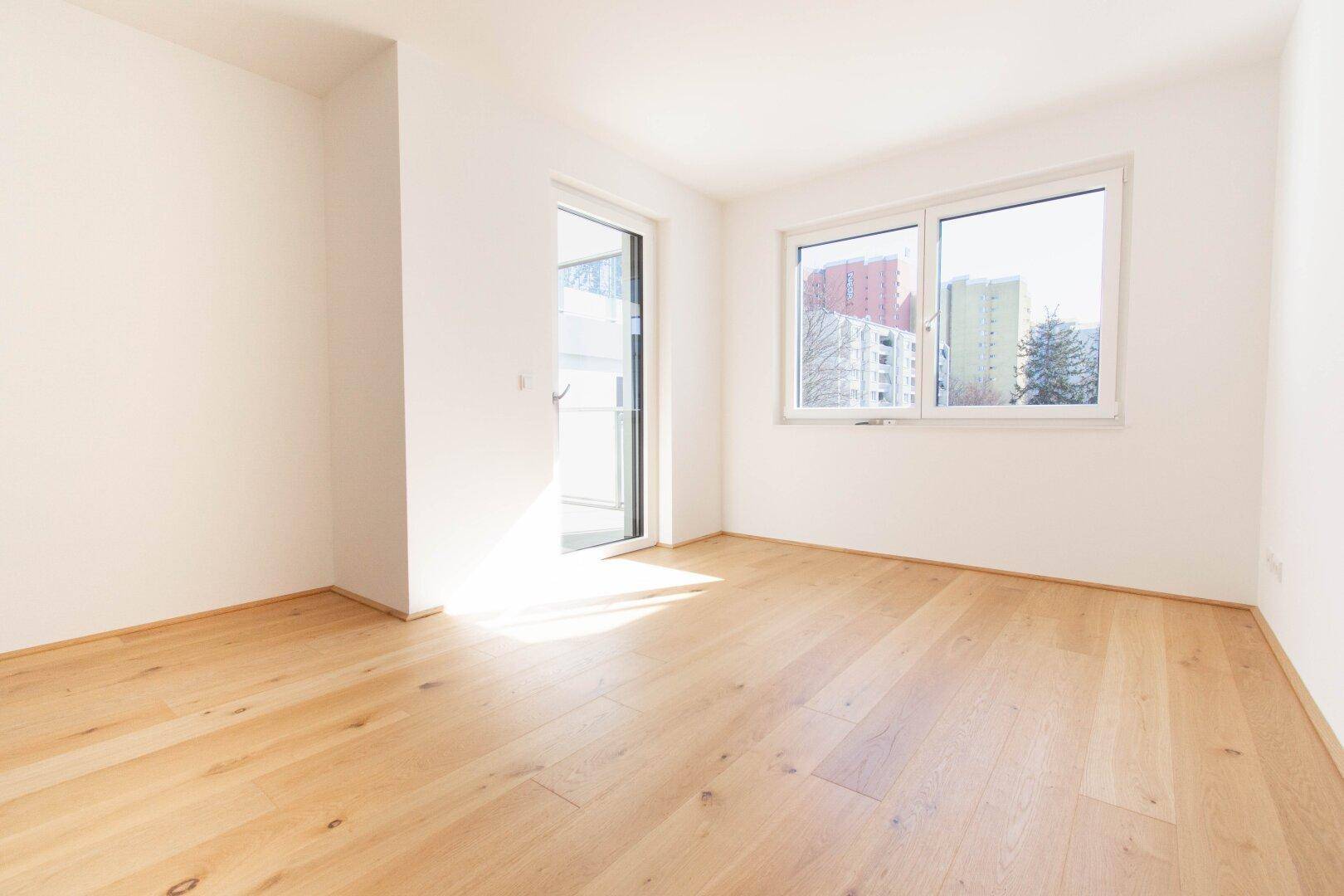 Geräumiges Zimmer mit Holzboden und großen Fenstern, das viel Tageslicht hereinlässt.
