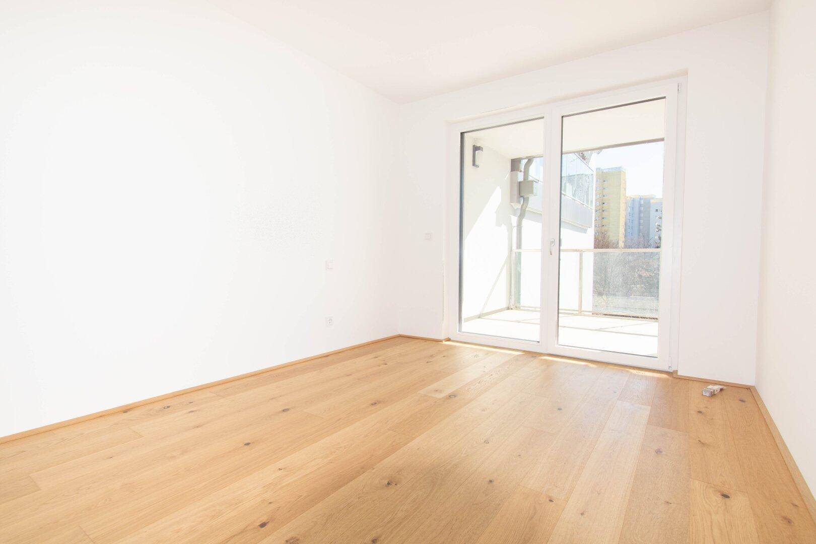 Lichtdurchfluteter Raum mit Holzboden und Zugang zu einem kleinen Balkon, ideal als Schlaf- oder Arbeitszimmer.
