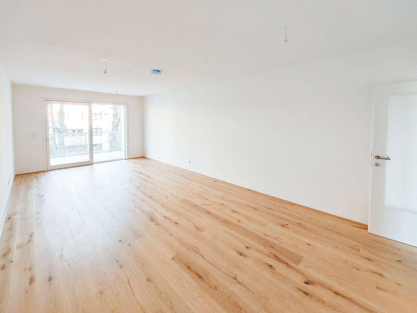Großzügiger Wohnbereich mit Holzboden und Zugang zu einem Balkon, bietet viel Gestaltungsfreiheit.