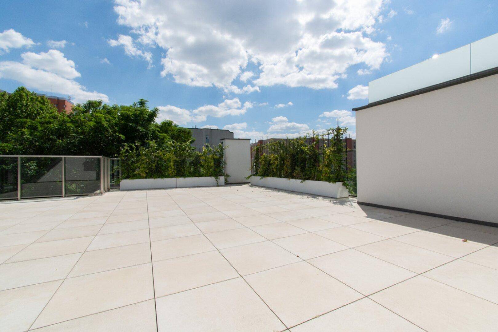 Weitläufige Dachterrasse mit Fliesenboden und begrünten Pflanzkübeln unter blauem Himmel.