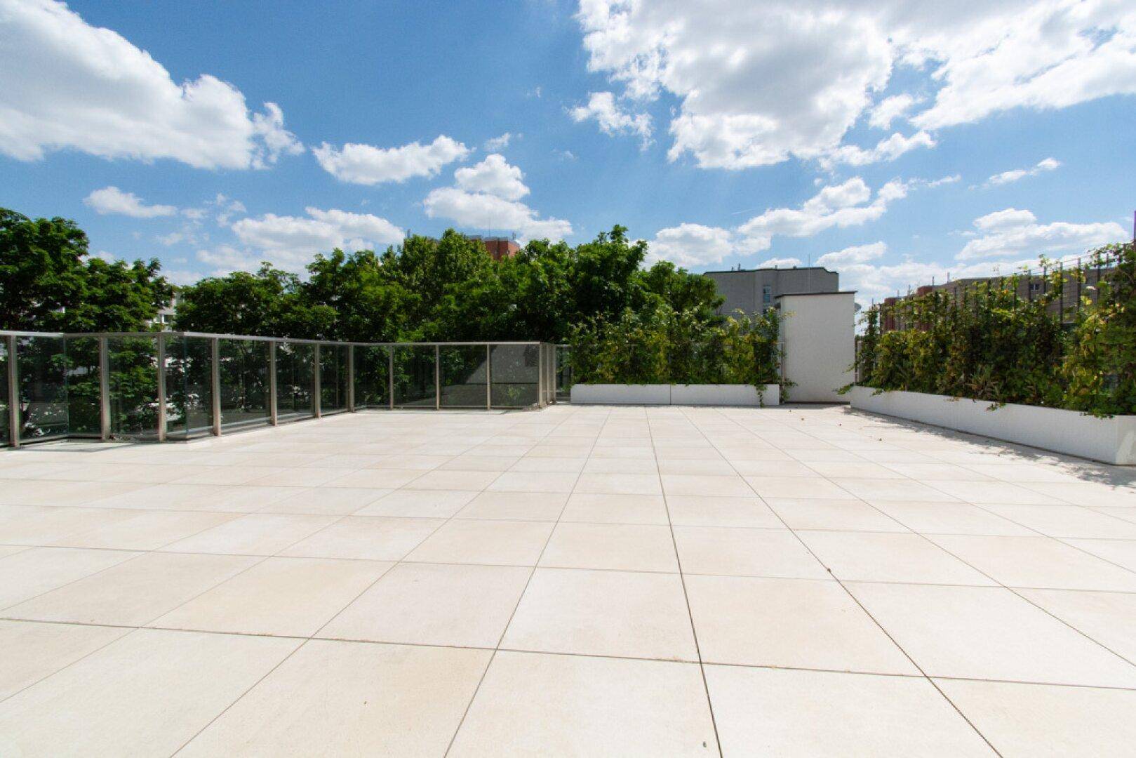 Geräumige Dachterrasse mit modernen Fliesen und Glasgeländer unter blauem Himmel.