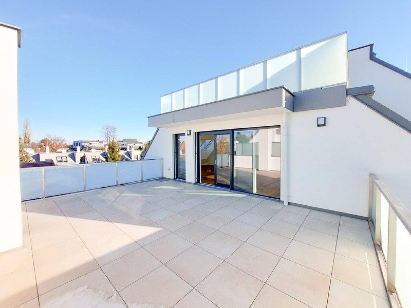 Geräumige Dachterrasse mit hellen Fliesen, Glasgeländer und weitem Blick unter blauem Himmel.