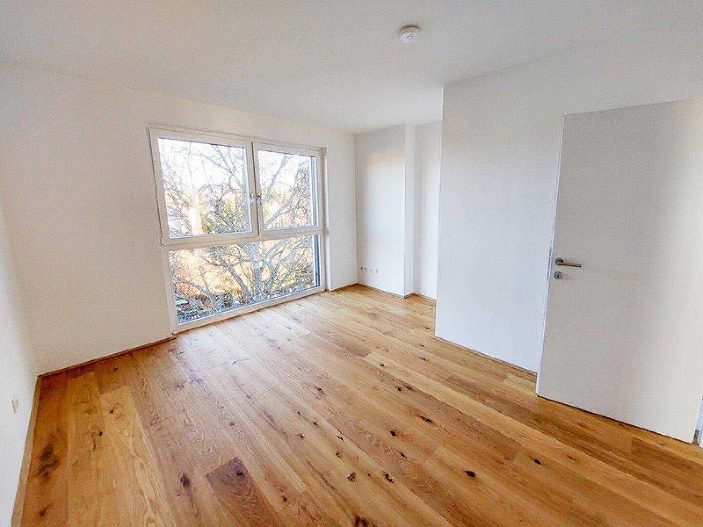 Leeres Schlafzimmer mit Holzboden und großem Fenster, das einen Blick ins Grüne bietet.