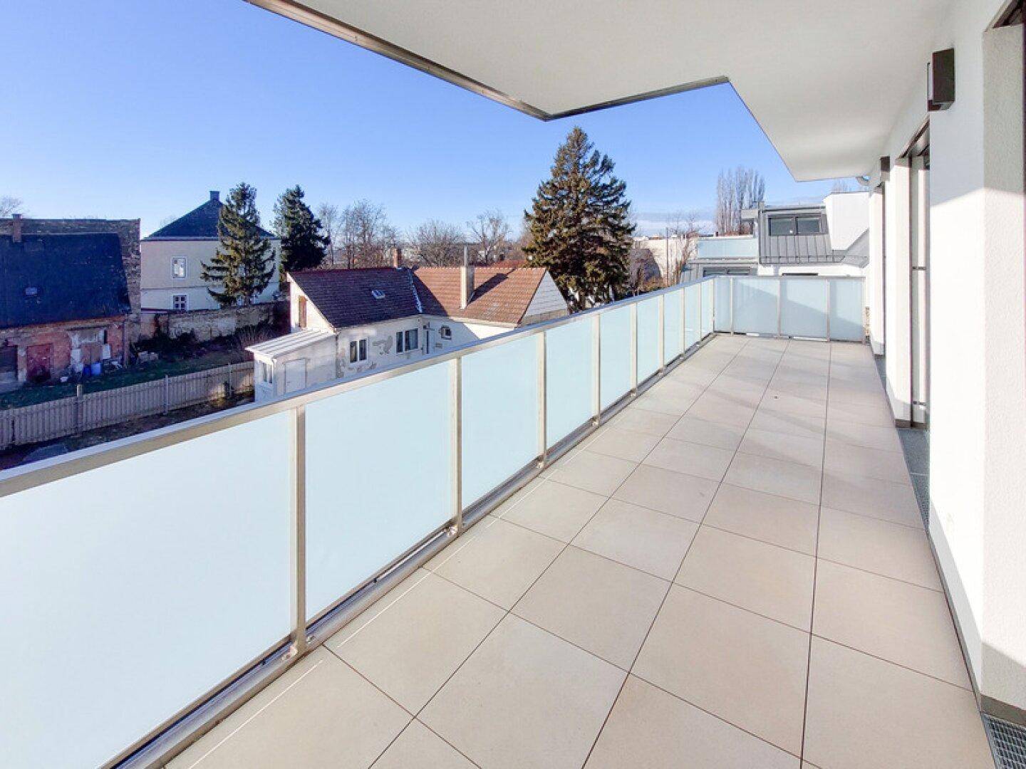 Langer Balkon mit hellen Fliesen und Milchglasgeländer, bietet Blick auf die umliegenden Häuser.
