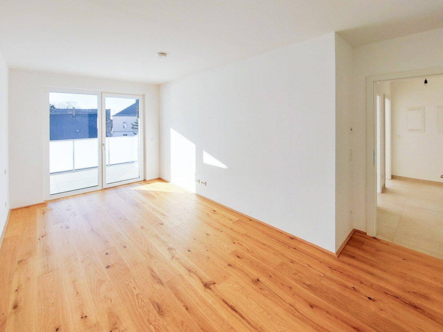 Heller, leerer Wohnraum mit Holzboden und Zugang zu einem Balkon oder einer Terrasse.