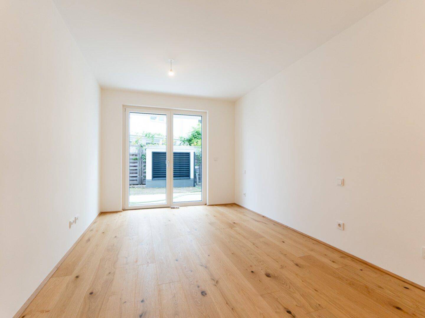 Helles Zimmer mit Holzboden und direktem Zugang zum Gartenbereich durch eine Glastür.