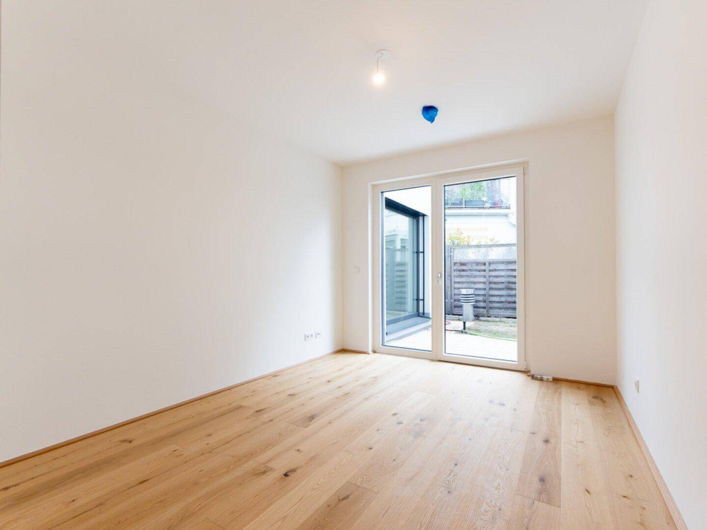 Leeres Zimmer mit Holzboden und Zugang zu einem kleinen Außenbereich durch eine Glastür.