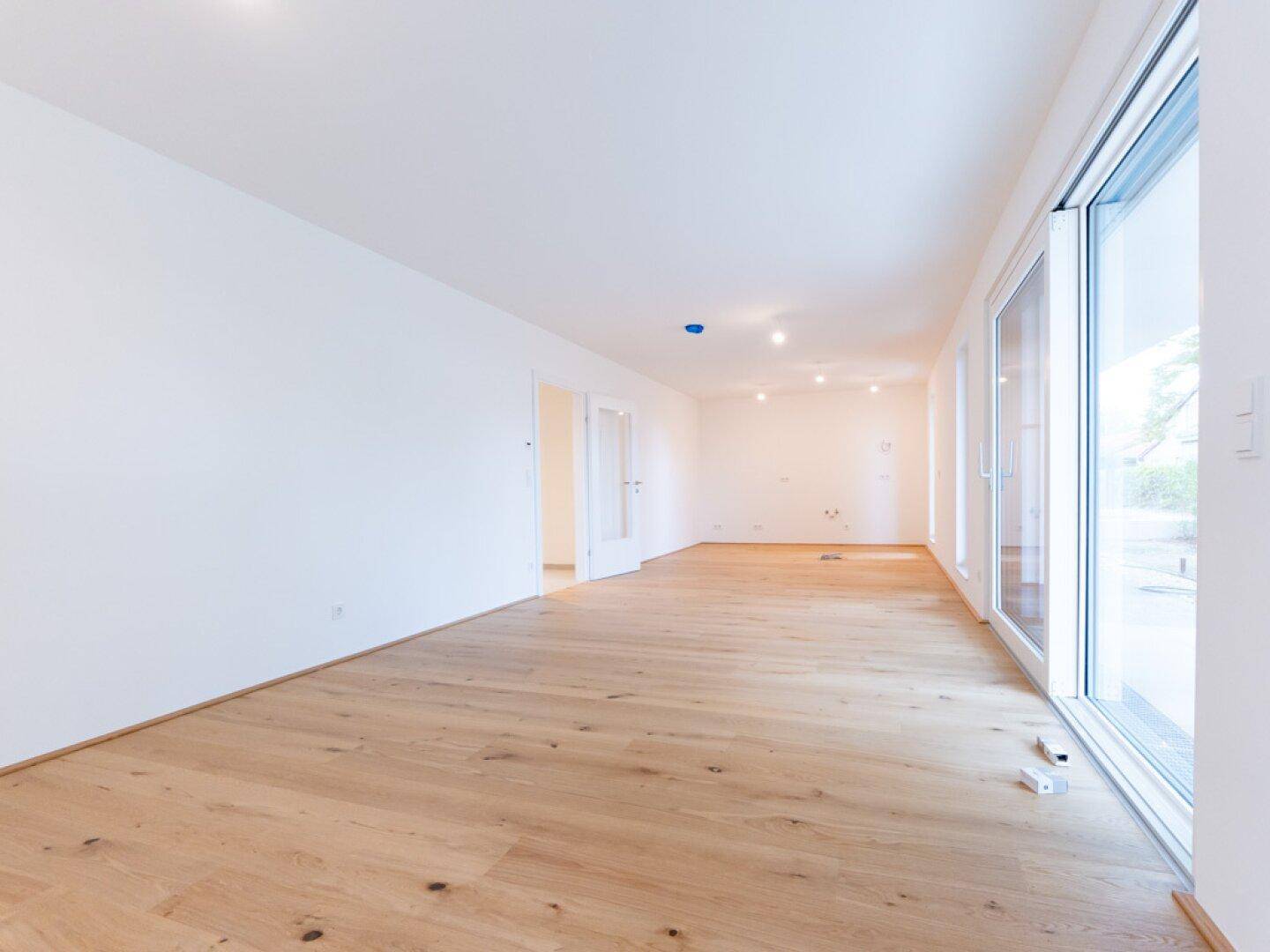 Geräumiger, heller Raum mit Holzboden und bodentiefen Fenstern, ideal für einen Wohnbereich.