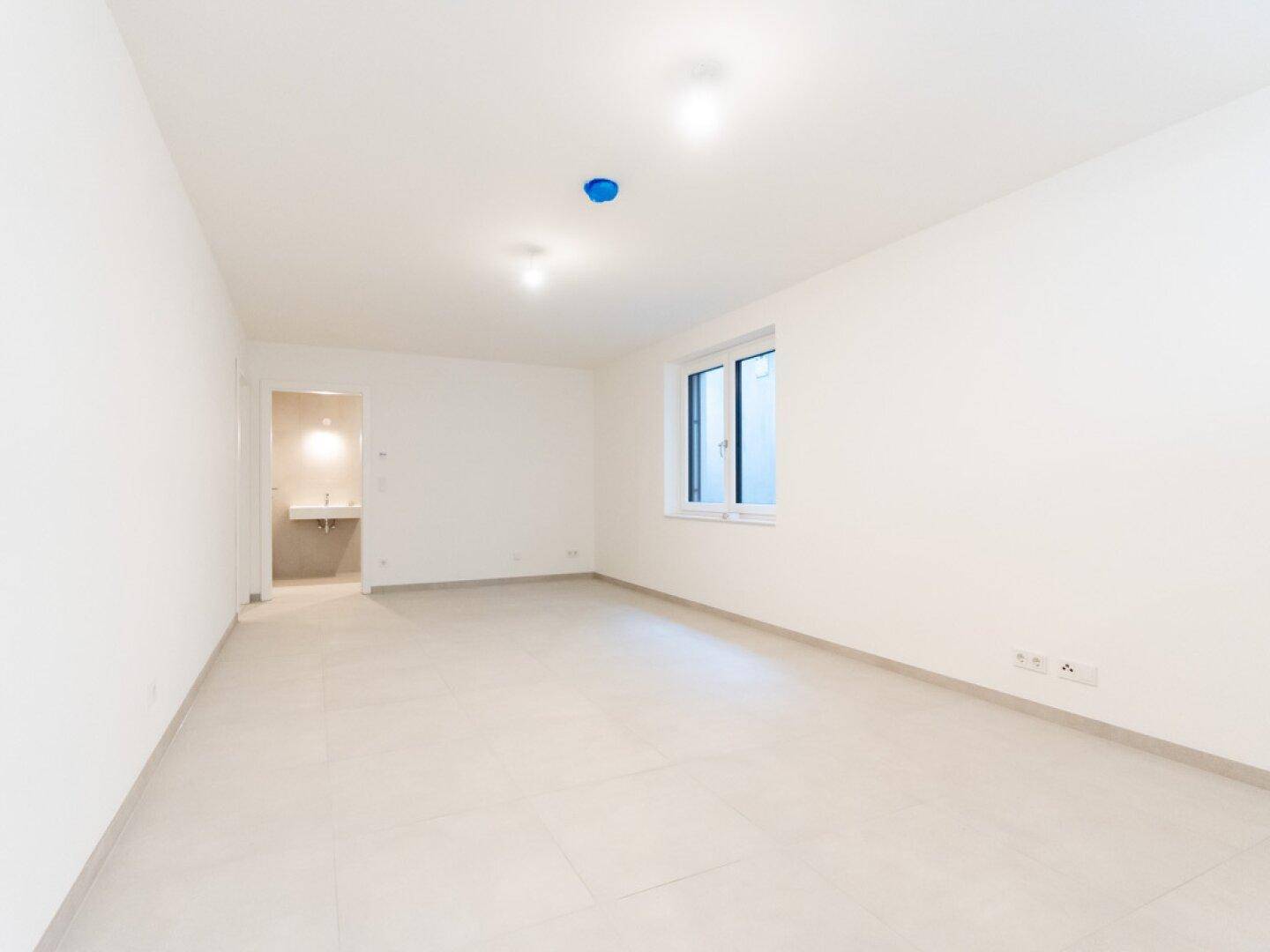 Geräumiger, heller Raum mit Fliesenboden und Fenster, angrenzend an einen Waschraum.