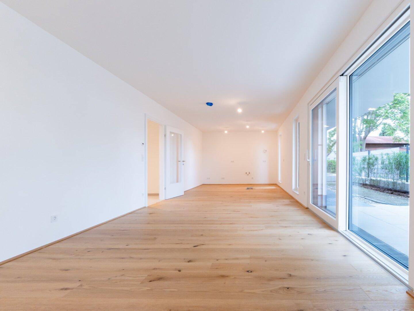 Heller, leerer Raum mit Holzboden und großen Fenstern, die viel Tageslicht hereinlassen.