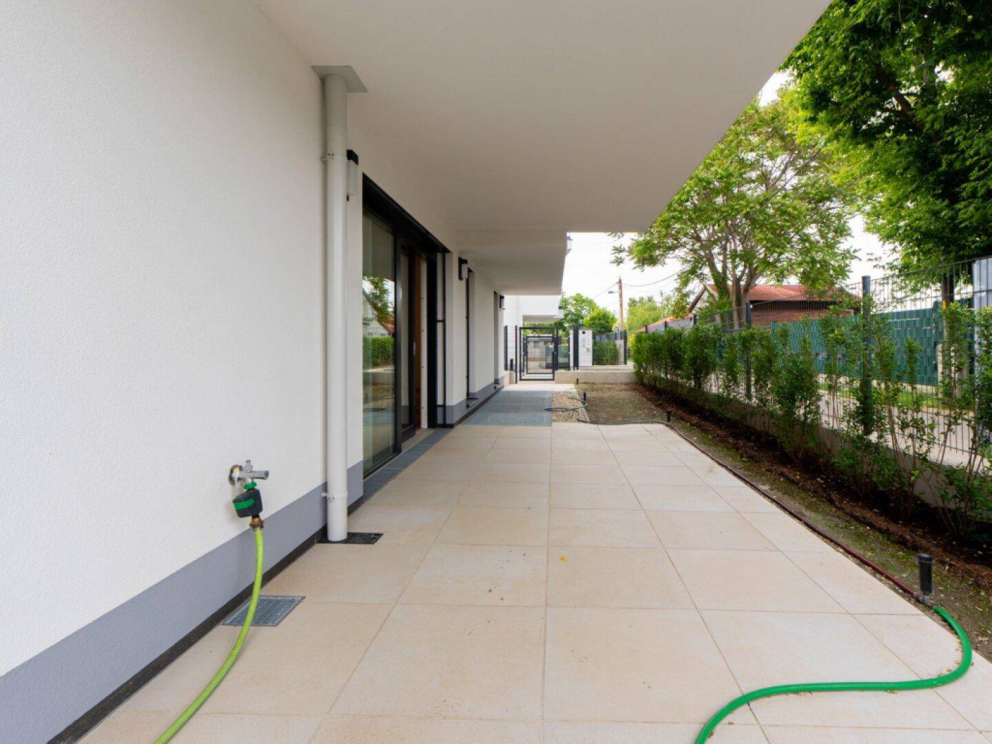 Gepflasterte Terrasse entlang des Gebäudes mit Wasseranschluss und Hecke im Hintergrund.