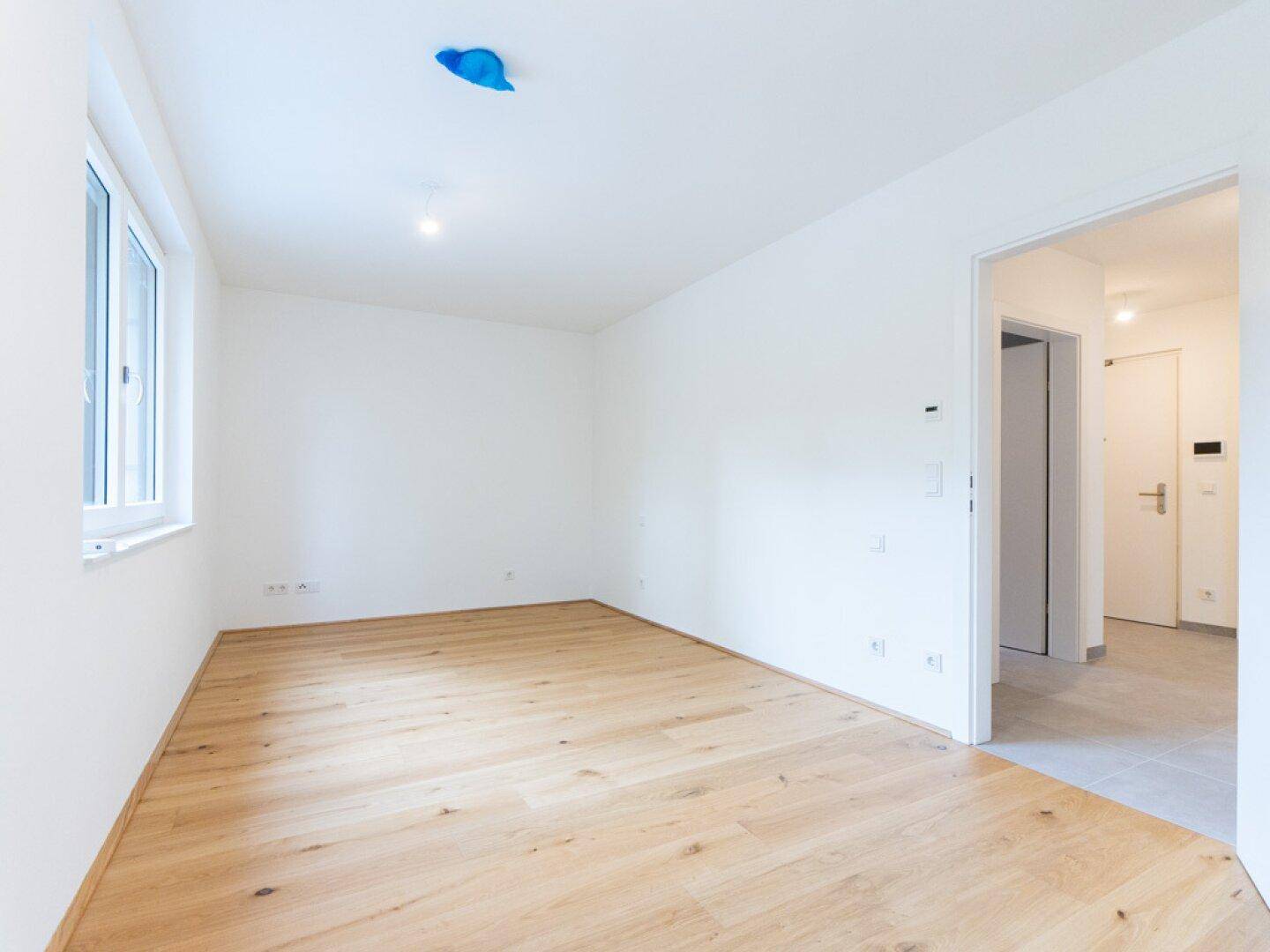 Freundliches Zimmer mit hellem Holzparkett und Fenster, angrenzend an einen gefliesten Flur.