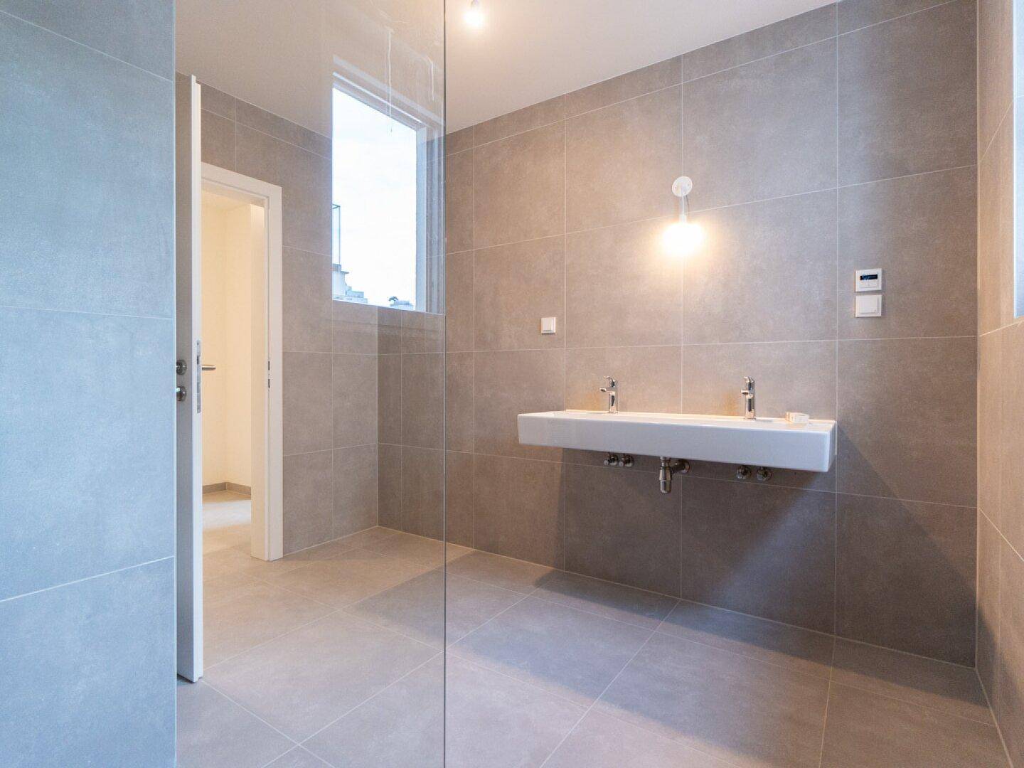 Modernes Badezimmer mit grauen Fliesen, Doppelwaschbecken und einem Fenster für natürliche Belüftung.