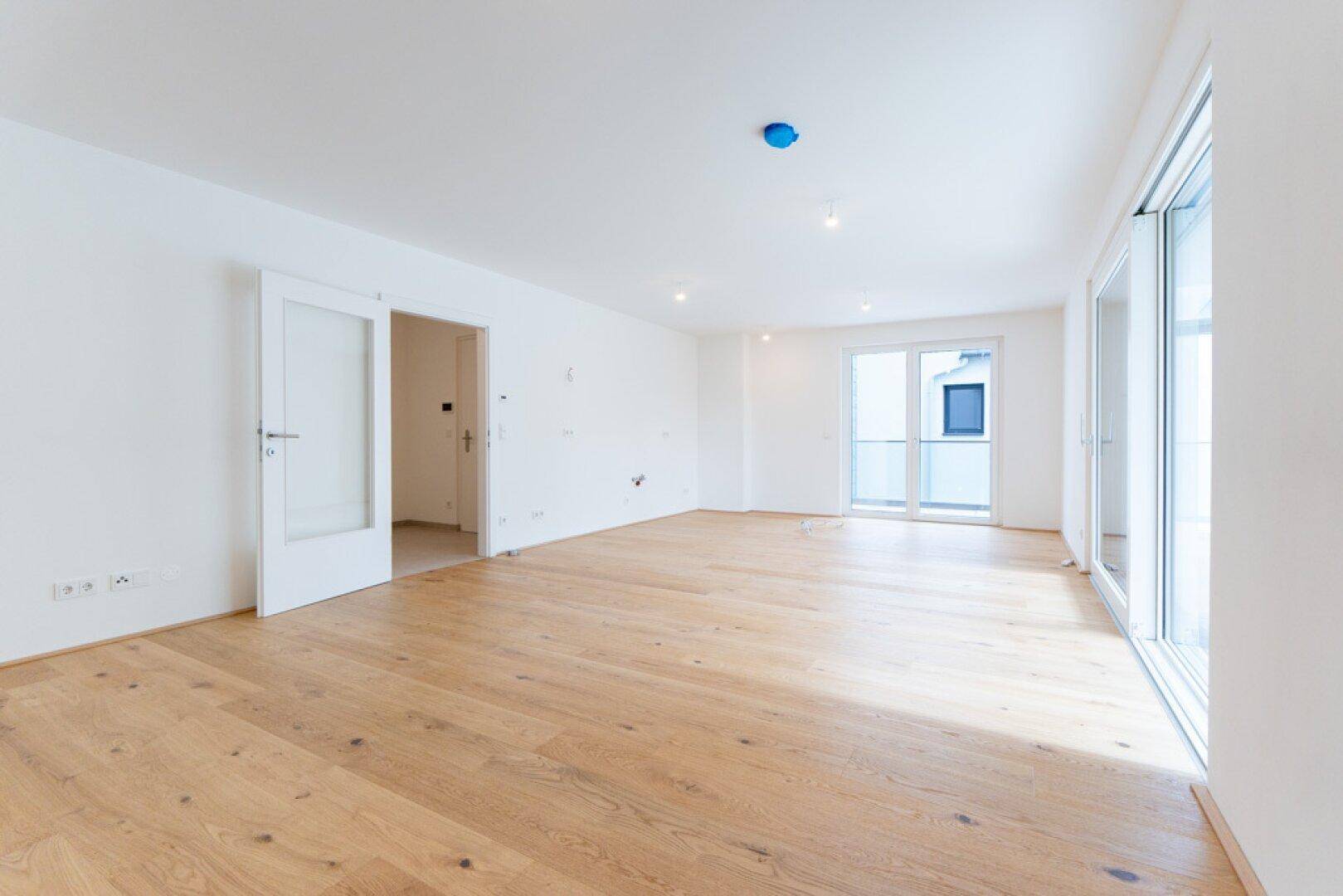 Heller Wohnraum mit Holzboden, großen Fenstern und Zugang zu einem kleinen Balkon.