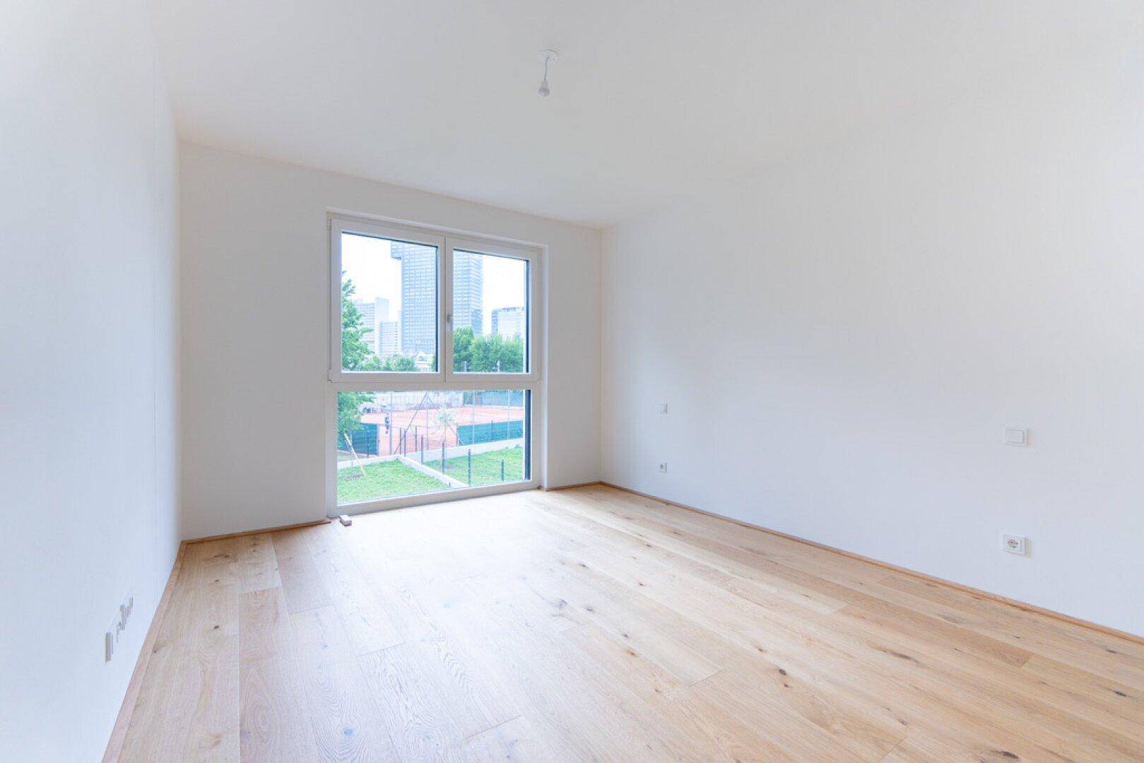 Helles Zimmer mit Holzboden und großem Fenster mit Blick auf den Tennisplatz und die Stadt.