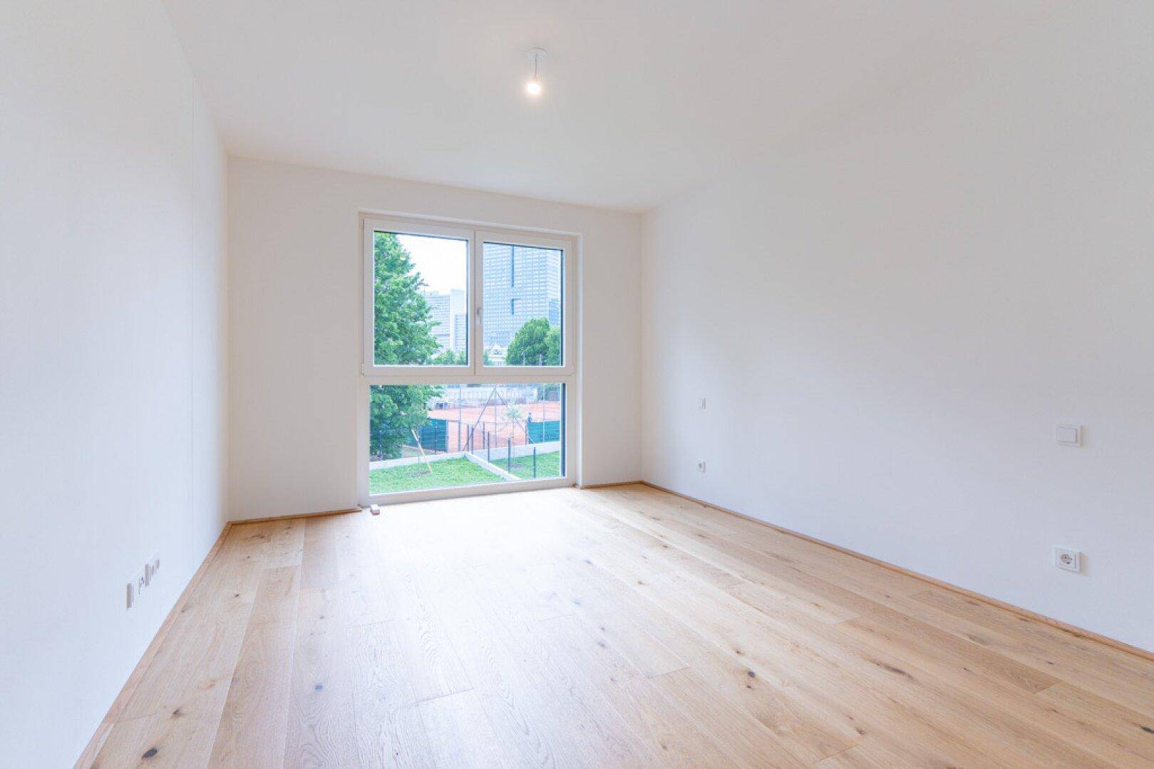Helles Zimmer mit Holzboden und großem Fenster mit Blick auf den Tennisplatz und die Stadt.