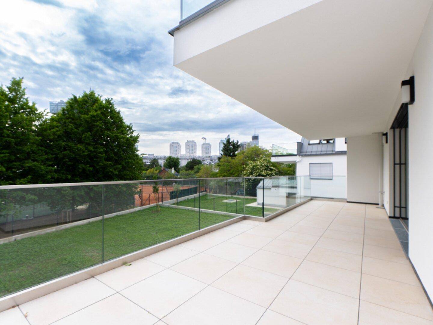 Weitläufige Terrasse mit Fliesenboden, Glasgeländer und Blick auf Tennisplatz und Stadt.