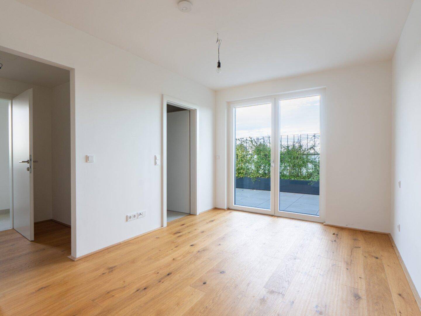 Heller, leerer Raum mit Holzboden und Zugang zu einer begrünten Terrasse.