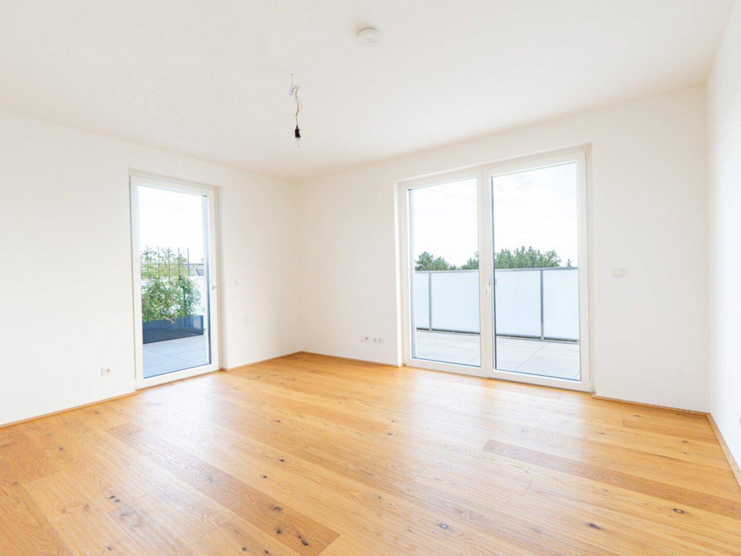 Leerer, lichtdurchfluteter Raum mit Holzboden und großen Fenstern, die auf eine Terrasse führen.