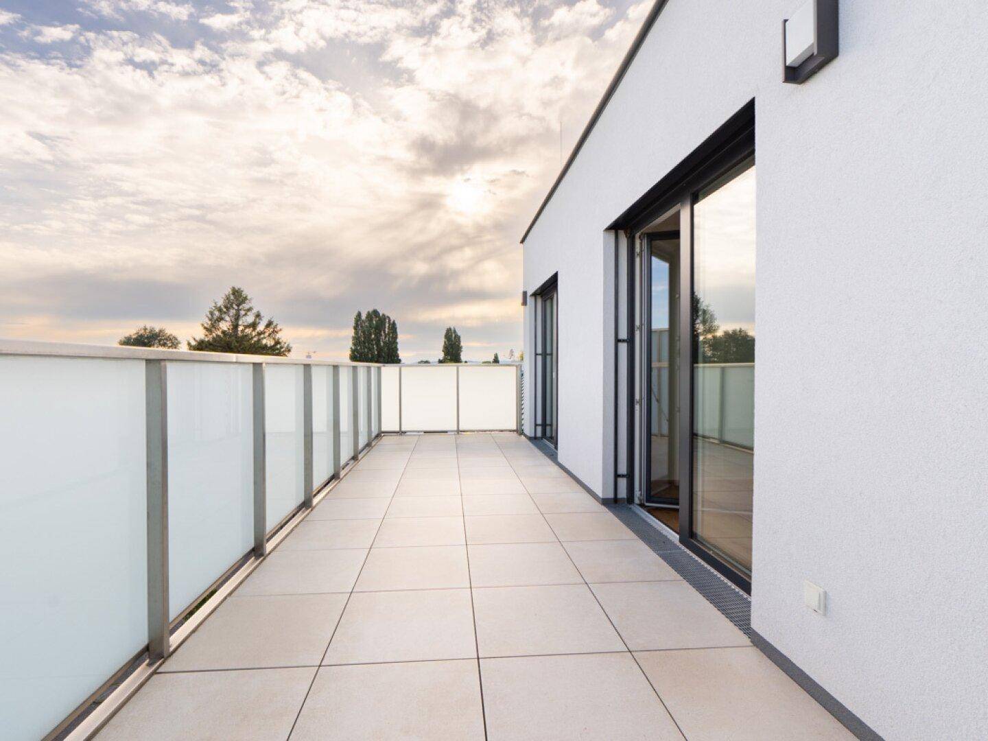 Moderne, geflieste Terrasse mit Glasgeländer und weitem Blick unter einem bewölkten Himmel.