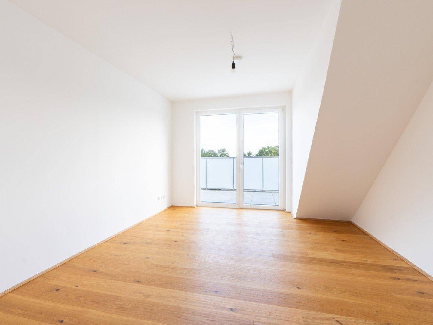 Heller, leerer Raum mit Holzboden, Dachschräge und Zugang zu einer sonnigen Terrasse.
