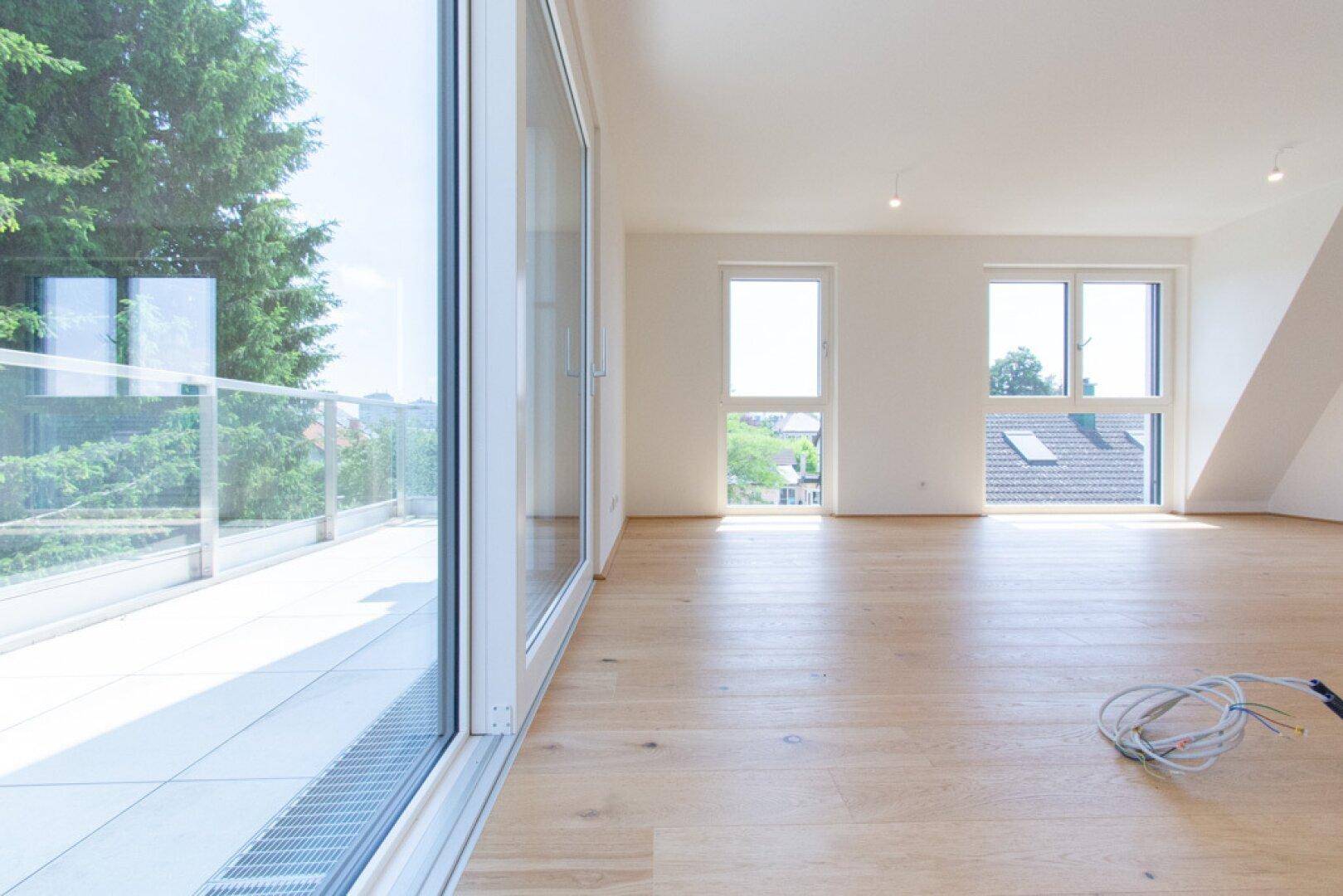 Heller, unmöblierter Wohnbereich mit großen Fenstern, Holzboden und Zugang zum Balkon.