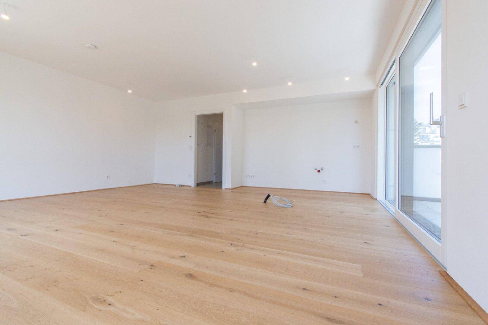 Geräumiger, leerer Wohnbereich mit hellem Parkettboden und Zugang zu einem Balkon.