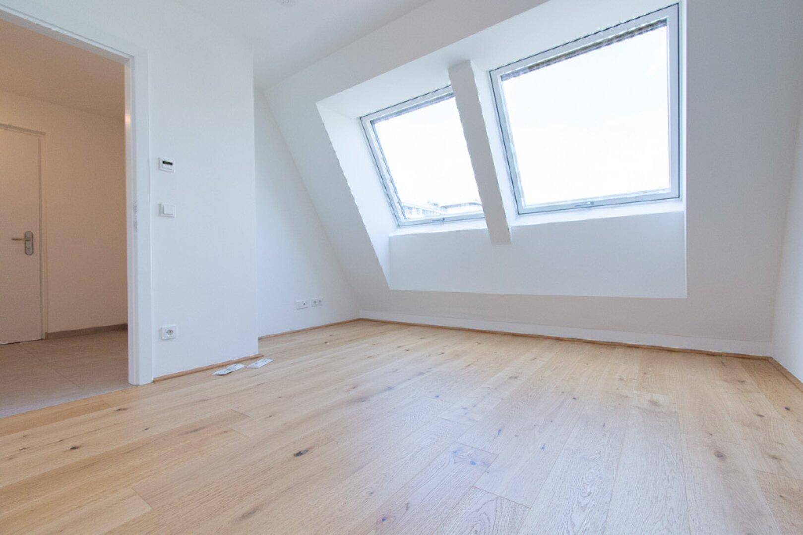 Helles Zimmer im Dachgeschoss mit schrägen Wänden, Holzboden und zwei Dachfenstern.