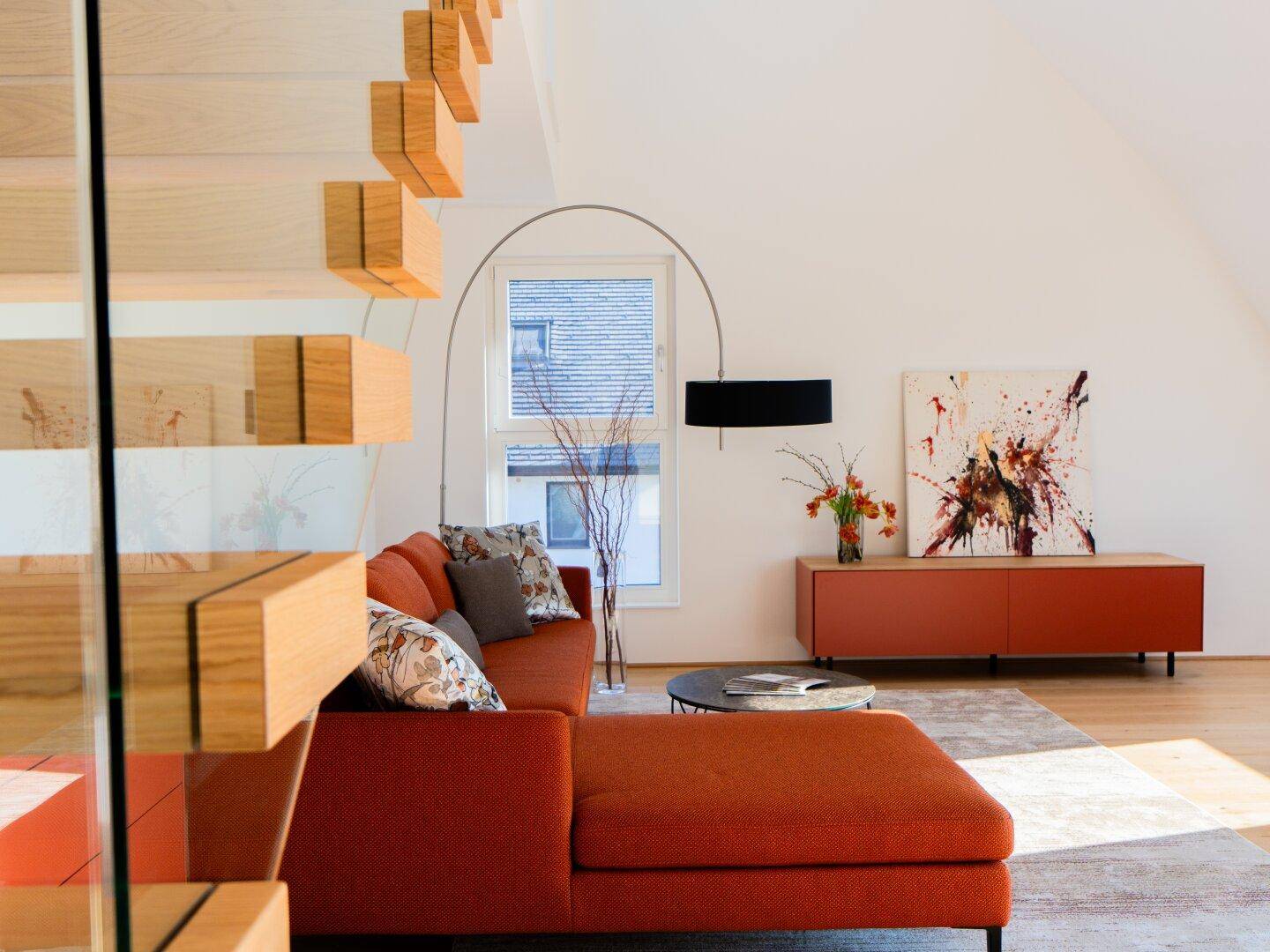 Moderner Wohnbereich mit rotem Sofa, Bogenlampe und offener Holztreppe, die den Raum prägt.