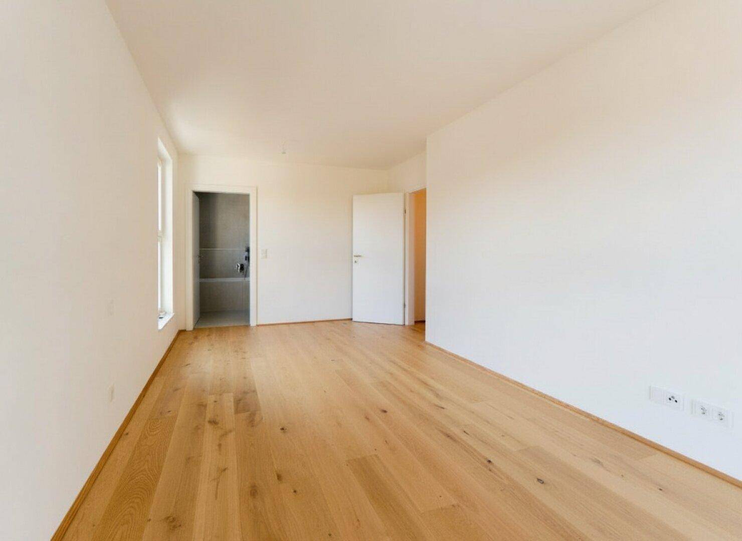 Leeres Schlafzimmer mit hellem Parkettboden und Zugang zu einem angrenzenden Badezimmer.