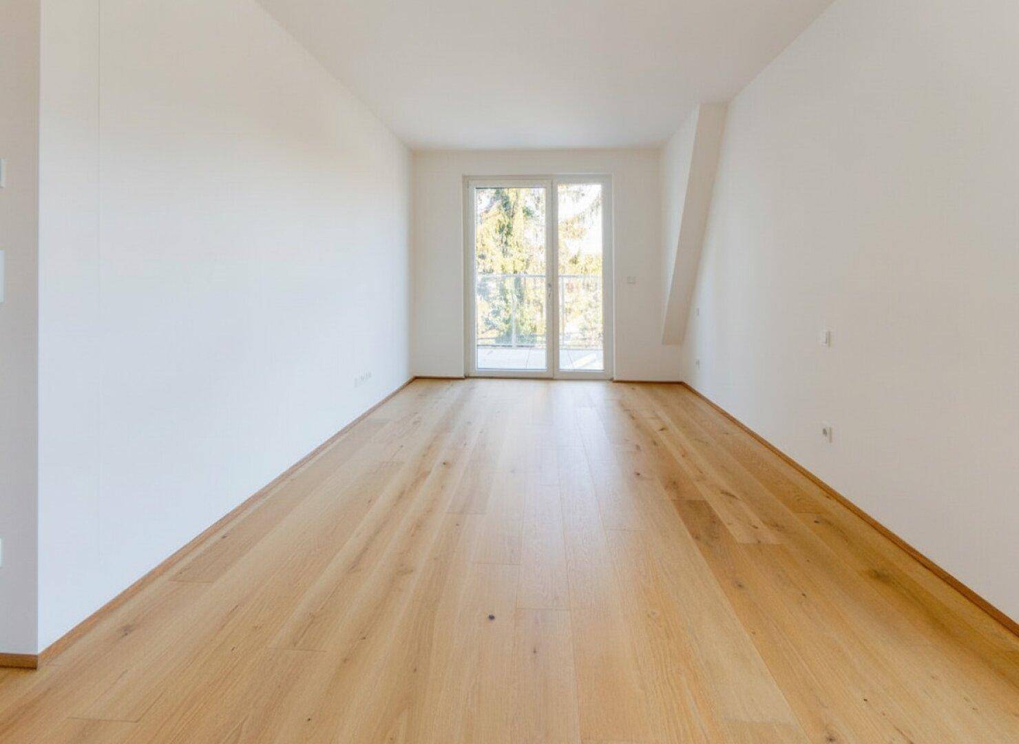 Leerer Raum mit hellem Holzboden und französischem Balkon, der viel Licht hereinlässt.