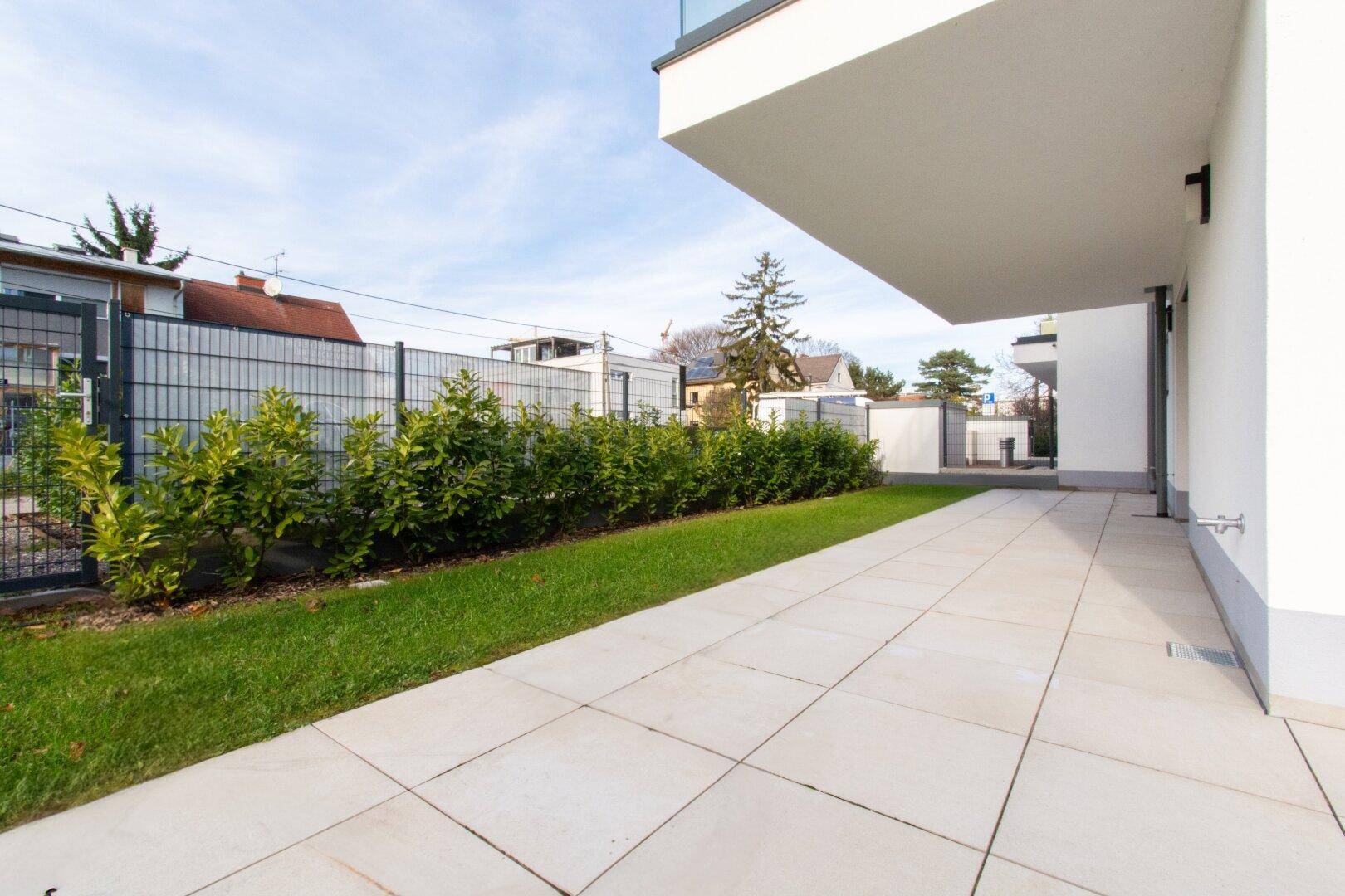 Lange, gepflasterte Terrasse mit Rasenstreifen und Hecke entlang der Grundstücksgrenze.