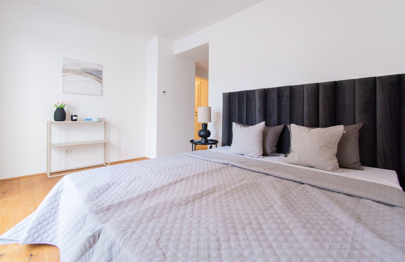 Schlafzimmer mit großem Bett, dunklem Kopfteil und minimalistischer Dekoration.