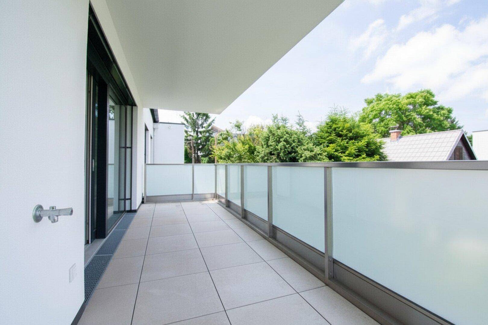 Moderner Balkon mit Glasbrüstung und Fliesenboden, umgeben von grüner Vegetation und Bäumen.