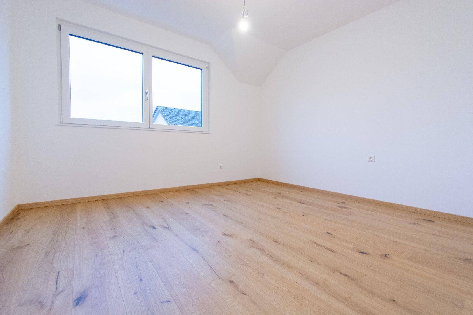 Helles Schlafzimmer im Dachgeschoss mit Holzparkettboden, weiß gestrichenen Wänden und einem Fenster.