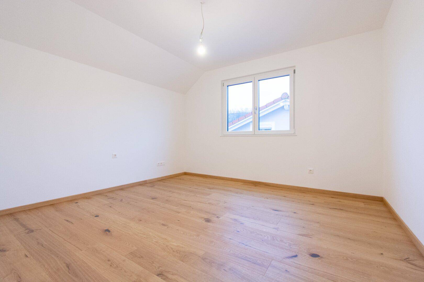 Dachgeschosszimmer mit Holzparkettboden, weiß gestrichenen Wänden und einem Fenster mit Blick nach draußen.