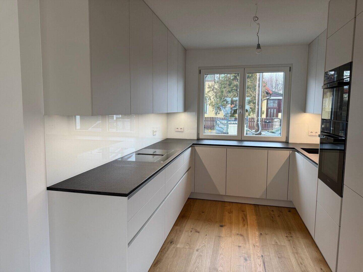 Moderne Einbauküche in Weiß mit dunkler Arbeitsplatte, integrierten Geräten und Fensterblick nach draußen.